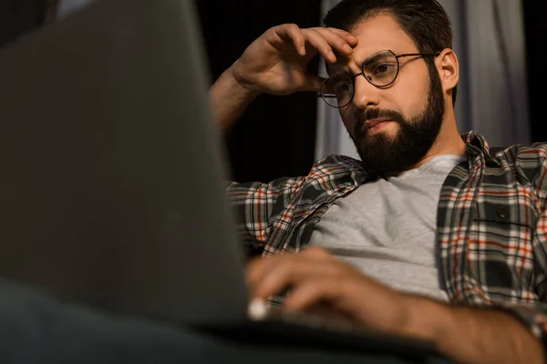 Homem cansado em óculos sentado no sofá com laptop — Fotografia de Stock