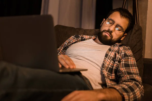 Homme fatigué dans des lunettes assis sur le canapé avec ordinateur portable — Photo de stock
