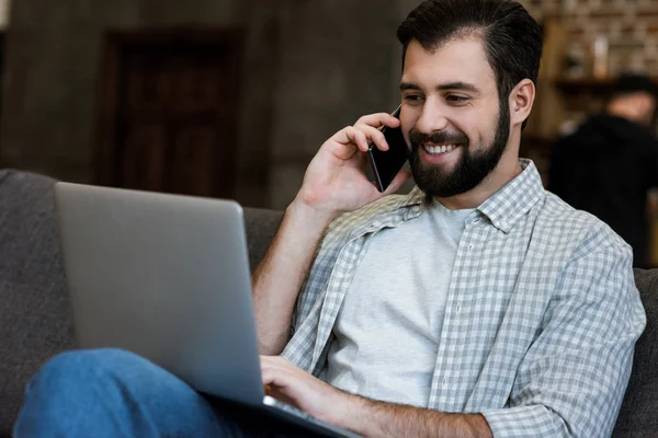 Hombre guapo sentado en el sofá con el ordenador portátil y hablando por teléfono - foto de stock