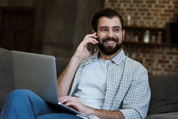 Handsome man sitting on couch with laptop and speaking on phone — Stock Photo