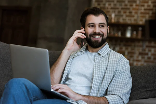 Hombre guapo sentado en el sofá con el ordenador portátil y hablando por teléfono - foto de stock