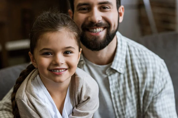 Fröhlicher Vater und Tochter, die sich umarmen und zu Hause in die Kamera schauen — Stockfoto