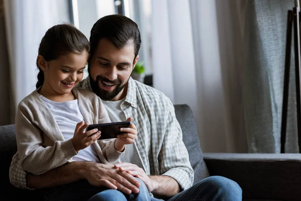 Padre alegre con hija abrazándose en el sofá y usando el teléfono inteligente en casa - foto de stock