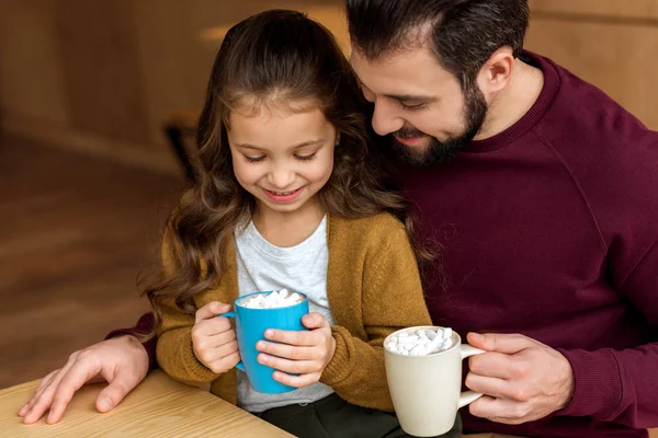 Entzückende Tochter sitzt auf den Knien des Vaters und hält eine Tasse Kakao mit Marshmallow in der Hand und blickt nach unten — Stockfoto