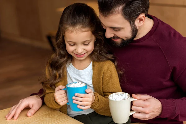 Adorable hija sentado en padre rodillas y sosteniendo taza de cacao con malvavisco y mirando hacia abajo - foto de stock