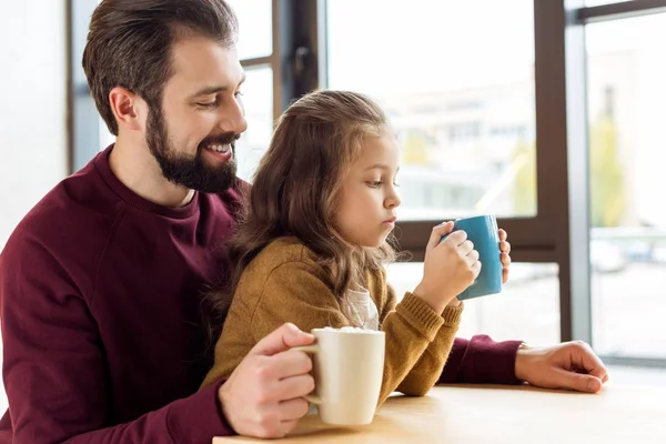 Adorable fille assise sur les genoux du père et tenant une tasse de cacao avec guimauve — Photo de stock