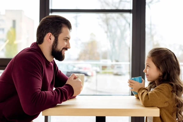 Feliz padre e hija sosteniendo tazas en la cafetería y mirándose - foto de stock