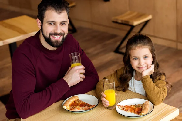 Счастливые отец и дочь пьют апельсиновый сок в кафе и смотрят в камеру — стоковое фото