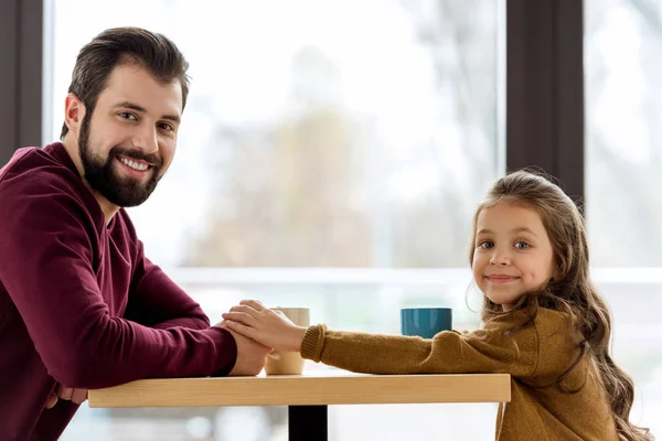 Sonrientes padre e hija sentados en la cafetería y tomados de la mano - foto de stock