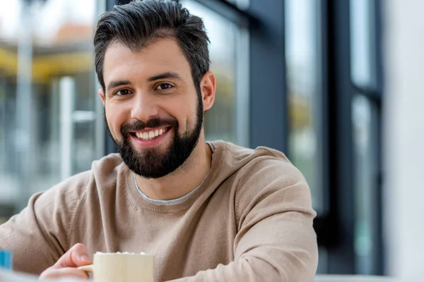 Smiling handsome man holding cup of coffee with marshmallow and looking at camera — Stock Photo