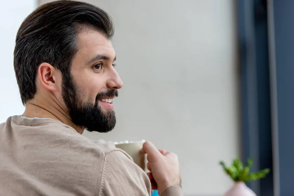 Sonriente hombre guapo sosteniendo taza de café con malvavisco y mirando hacia otro lado - foto de stock