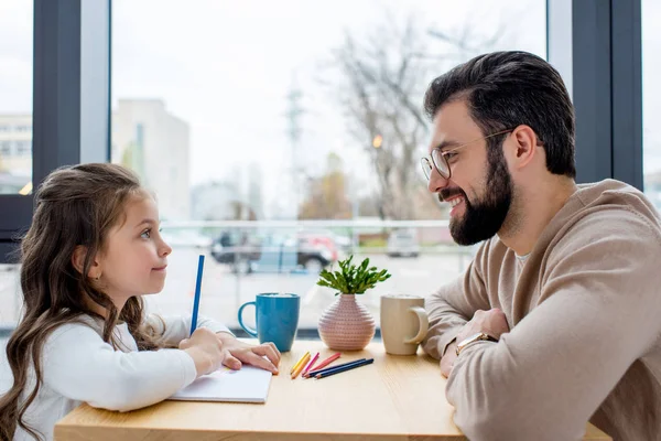 Smiling father looking at daughter while she drawing in album — Stock Photo