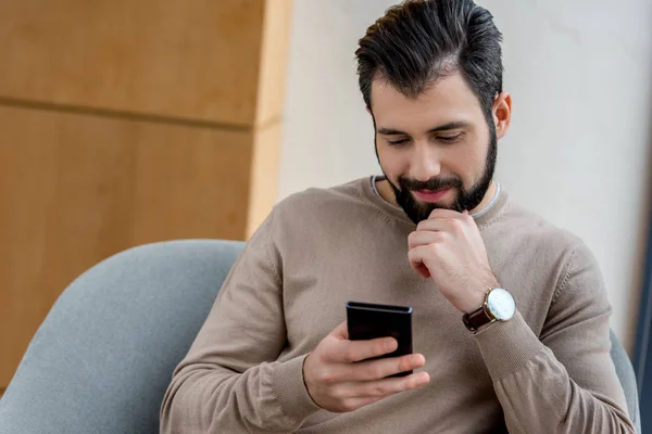 Handsome man sitting in armchair and looking at smartphone — Stock Photo