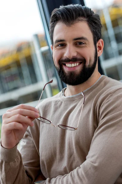 Retrato de hombre guapo sonriente sosteniendo gafas en la mano y mirando a la cámara - foto de stock