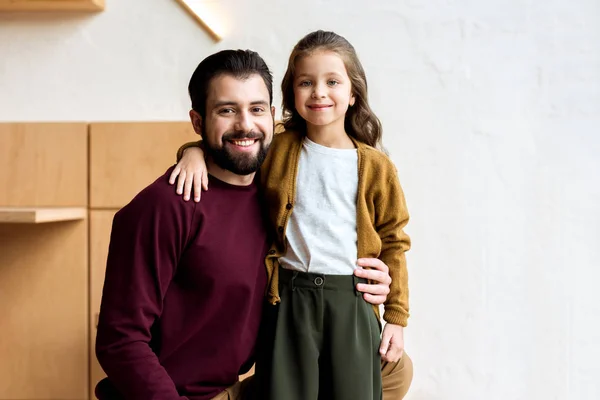 Padre en cuclillas y de pie hija abrazándose en la cafetería y mirando a la cámara - foto de stock