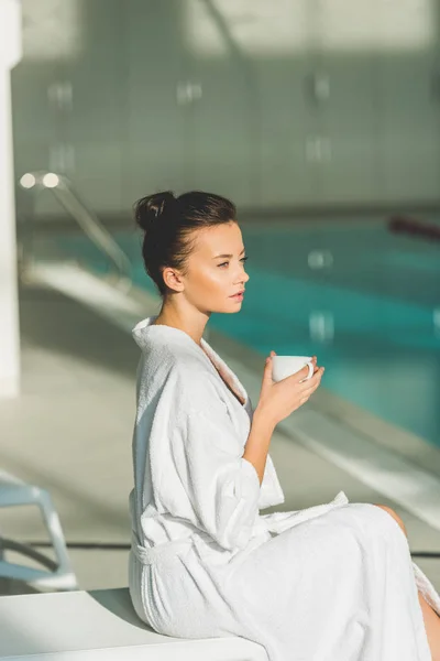 Schöne junge Frau im Bademantel mit Tasse Kaffee im Wellnessbereich — Stockfoto
