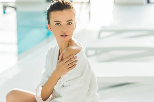 Beautiful young woman in bathrobe with naked shoulder touching her skin at spa — Stock Photo