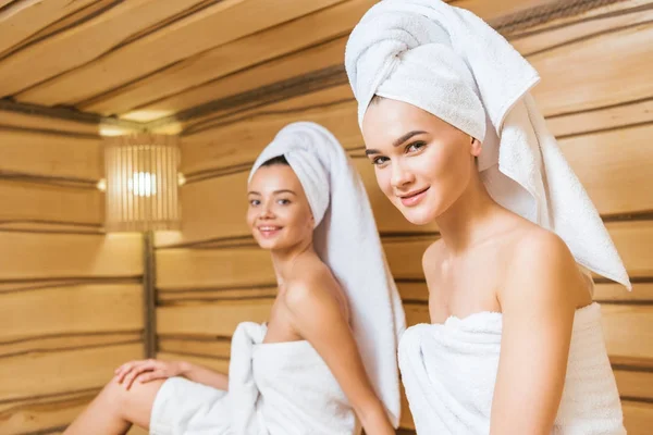 Mujeres jóvenes y felices cubiertas con toallas en la sauna y mirando a la cámara - foto de stock