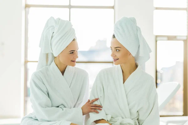 Hermosas mujeres jóvenes en albornoces y toallas en la cabeza hablando entre sí en el centro de spa - foto de stock