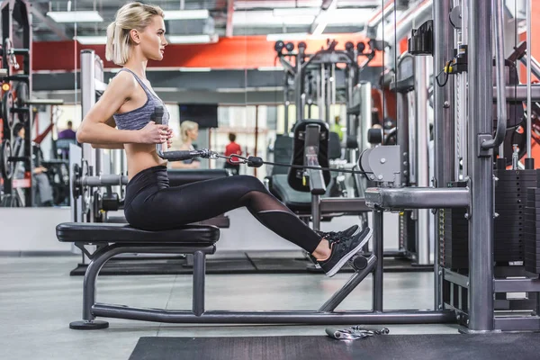 Vue latérale de la jeune femme travaillant avec la machine de rangée à la salle de gym — Photo de stock