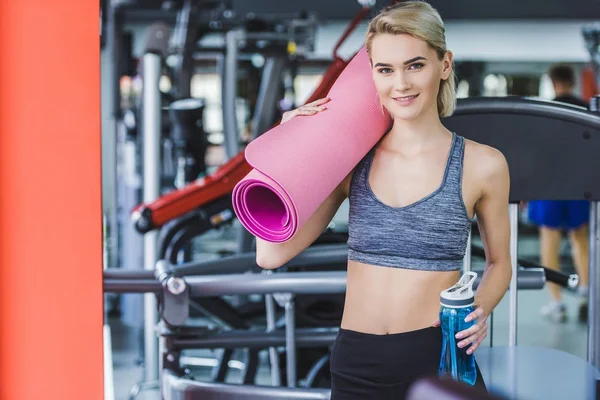 Atractiva joven con estera de yoga enrollada y botella de agua en el gimnasio - foto de stock