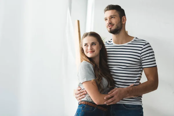 Young couple embracing near window and looking somewhere — Stock Photo