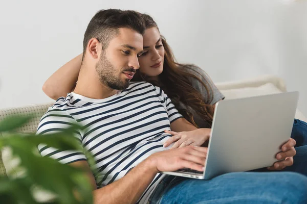 Mujer abrazando a su novio mientras él trabaja con el ordenador portátil en el sofá - foto de stock