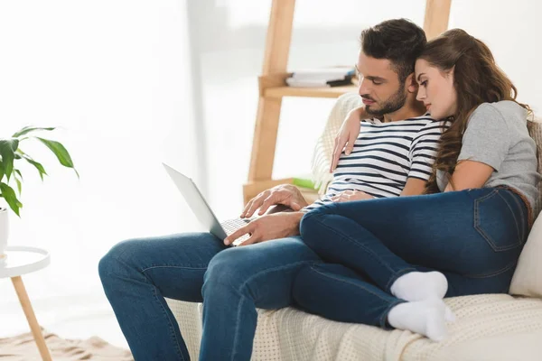 Cariñosa mujer abrazando a su novio mientras él trabaja con el ordenador portátil en casa - foto de stock