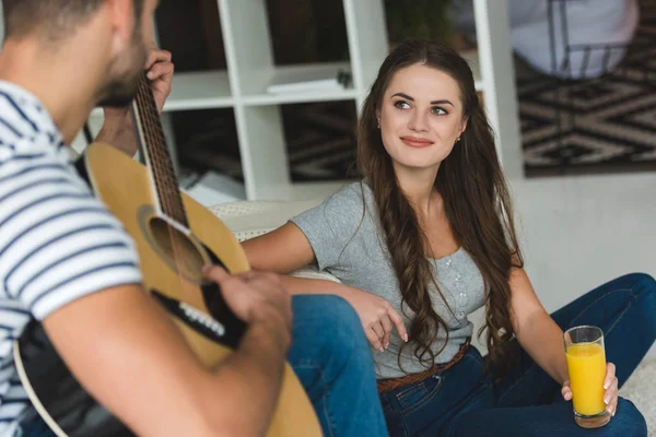 Hombre tocando la guitarra para su novia mientras ella está sentada en el suelo con jugo - foto de stock