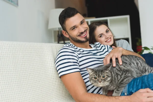 Belo jovem casal com adorável gato tabby sentado no sofá — Fotografia de Stock