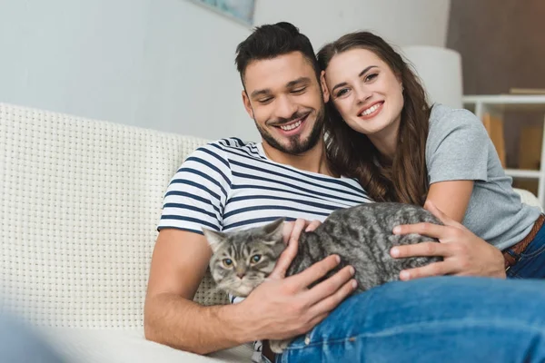 Glückliches junges Paar mit niedlicher gestromter Katze sitzt auf Couch — Stockfoto