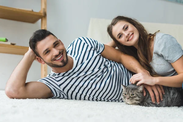 Beautiful young couple lying on floor with adorable tabby cat — Stock Photo