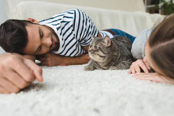 Feliz joven pareja acostada en el suelo con tabby gato - foto de stock