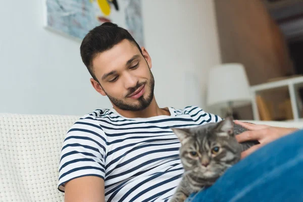 Schöner junger Mann streichelt süße gestromte Katze, während er auf der Couch sitzt — Stockfoto