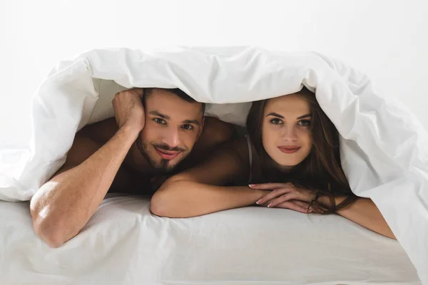 Jeune couple sous couverture regardant caméra dans le lit — Photo de stock