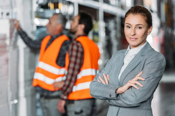 Ispettore donna pising con le braccia incrociate mentre gli uomini lavorano dietro nel magazzino — Foto stock