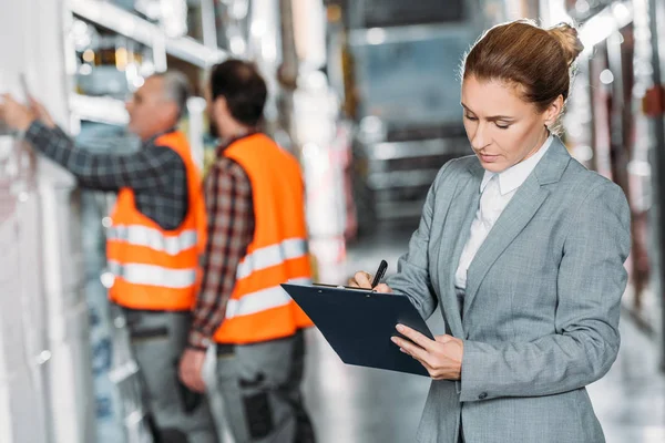 Female inspector writing something while men working behind in storehouse — Stock Photo