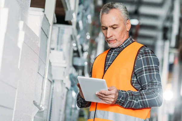 Senior worker in safety vest using digital tablet in storehouse — Stock Photo