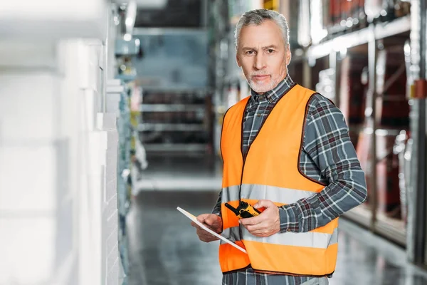 Senior worker in safety vest using digital tablet and walkie talkie in storehouse — Stock Photo