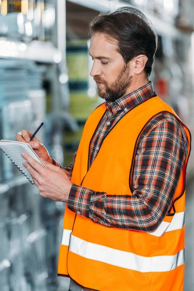 Male worker in safety vest writing in notepad in storehouse — Stock Photo