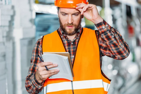 Nachdenklicher Arbeiter in Warnweste schaut auf Notizblock in Lagerhalle — Stockfoto