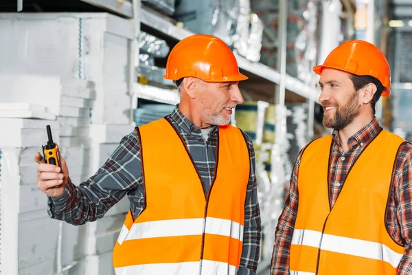 Two male workers in safety vests and helmets with walkie talkie in storehouse — Stock Photo