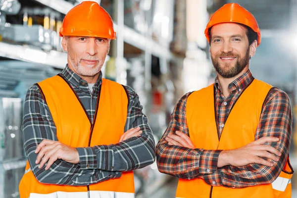 Two male workers in safety vests and helmets with crossed arms in storehouse — Stock Photo