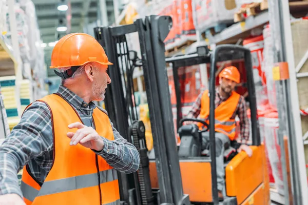 Trabajador masculino y su colega que trabajan con la máquina de la carretilla elevadora en almacén - foto de stock