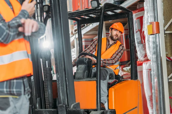 Worker in safety vest and helmet sitting in forklift machine in storage — Stock Photo