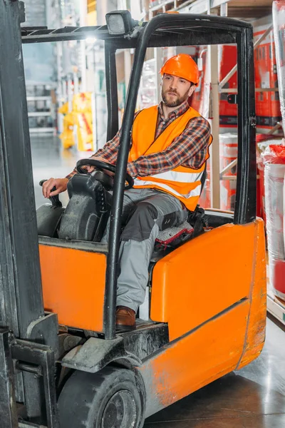 Male worker in safety vest and helmet sitting in forklift machine in storage — Stock Photo