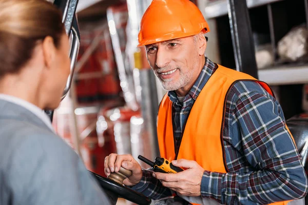 Senior worker with walkie talkie sitting in forklift machine and talking with businesswoman in storage — Stock Photo