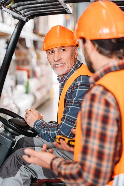 Senior worker in safety vest and helmet sitting in forklift machine and talking with colleague in storage — Stock Photo