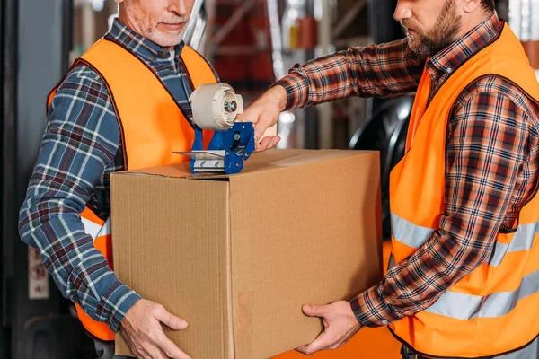 Cropped view of male workers in helmets packing cardboard box with scotch tape — Stock Photo