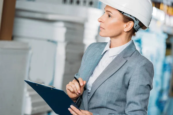 Female inspector in helmet writing notes on clipboard in warehouse — Stock Photo